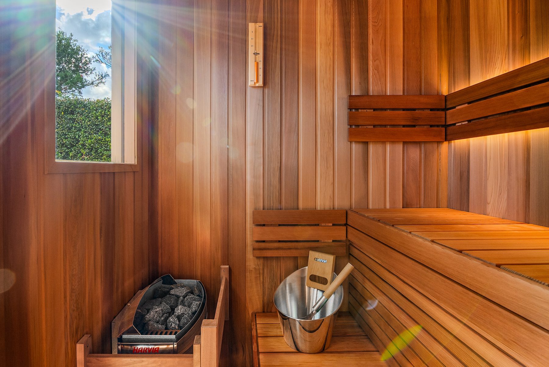 Western Red Cedar benches with sun shining in front window. Also pictured Harvia Vega heater and steel lux accessories set.